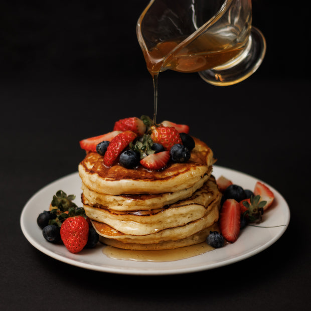 maple syrup pouring over a stack of pancakes strawberries and blueberries